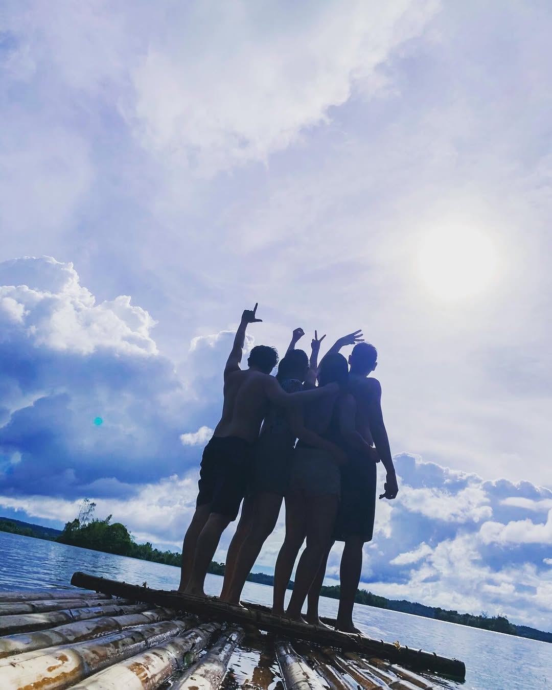 Silhouettes of four people on a bamboo raft cheering against a dramatic sky