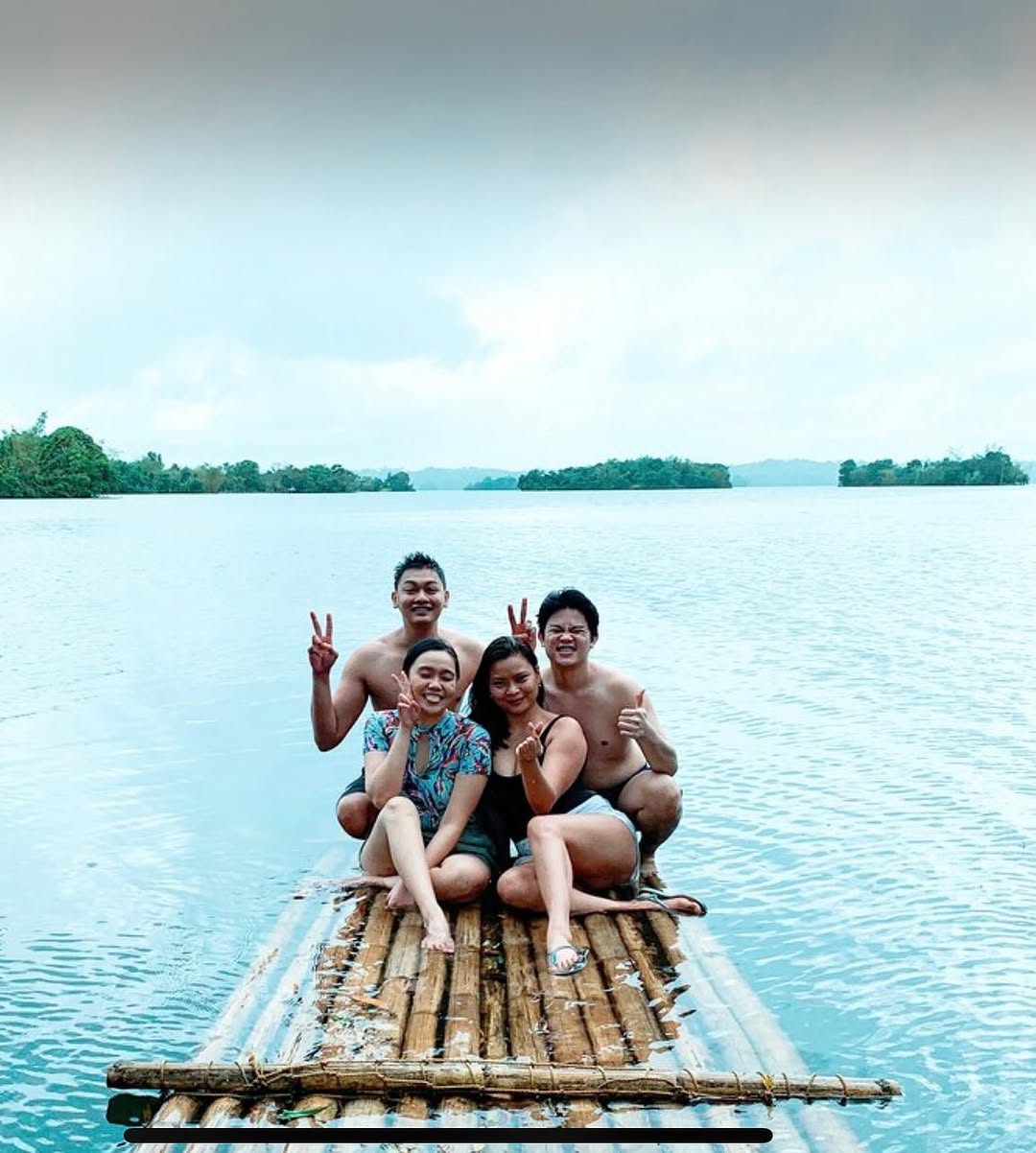 Four smiling guests posing on bamboo raft with vast lake and forest islands behind them