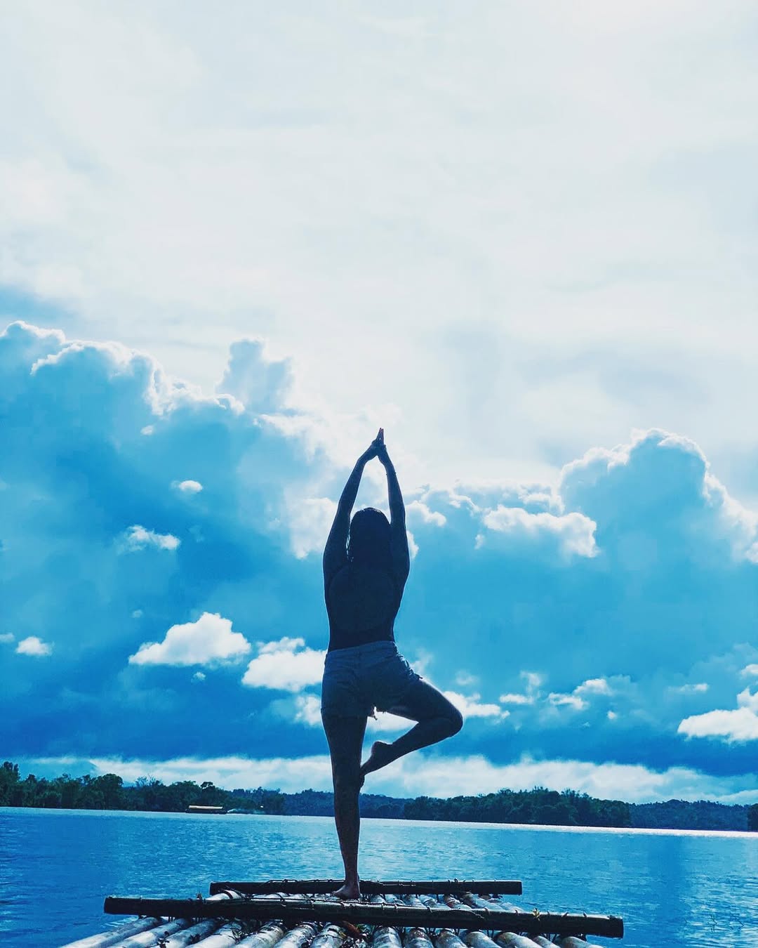 Person in yoga tree pose on bamboo raft, silhouetted against blue sky and clouds